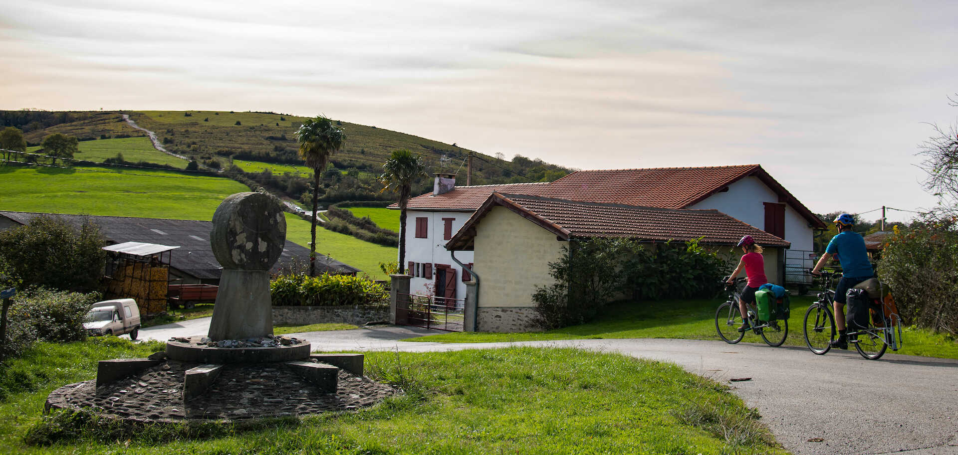 Des cyclistes passent devant un petit monument en pierre sur une route de campagne, avec une vue panoramique sur des collines verdoyantes et une maison en arrière-plan.